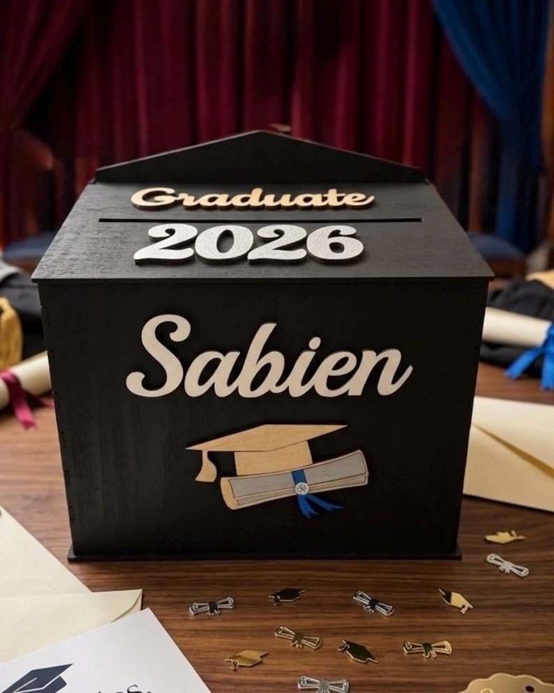 K&ouml;nnte beinhalten: Eine schwarze Holz-Abschlussbox mit den Worten "Graduate 2026" und "Sabien" in wei&szlig;er Schrift. Eine Abschlusskappe und ein Diplom sind ebenfalls abgebildet. Die Box steht auf einer Holzoberfl&auml;che mit Konfetti zum Thema Abschluss.