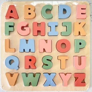 May include: A wooden alphabet puzzle with colorful letters in pastel shades of pink, blue, green, and orange. The letters are arranged in alphabetical order on a light brown wooden base.