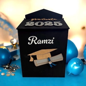 K&ouml;nnte beinhalten: Schwarze Abschlussbox mit dem Namen "Ramzi" in wei&szlig;er Schrift. Die Box hat ein Design mit Abschlusskappe und Diplom. Auf der Oberseite der Box steht "graduate 2025" in silbernen und goldenen Buchstaben. Blaue Luftballons und Konfetti im Hintergrund.