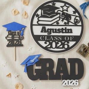 K&ouml;nnte beinhalten: Abschlussdekorationen, darunter ein gro&szlig;es schwarz-wei&szlig;es rundes Schild mit dem Namen "Agustin" und "Klasse von 2026". Au&szlig;erdem ein blau-schwarzes "Grad"-Schild mit "2026" und eine blaue Abschlusskappe.