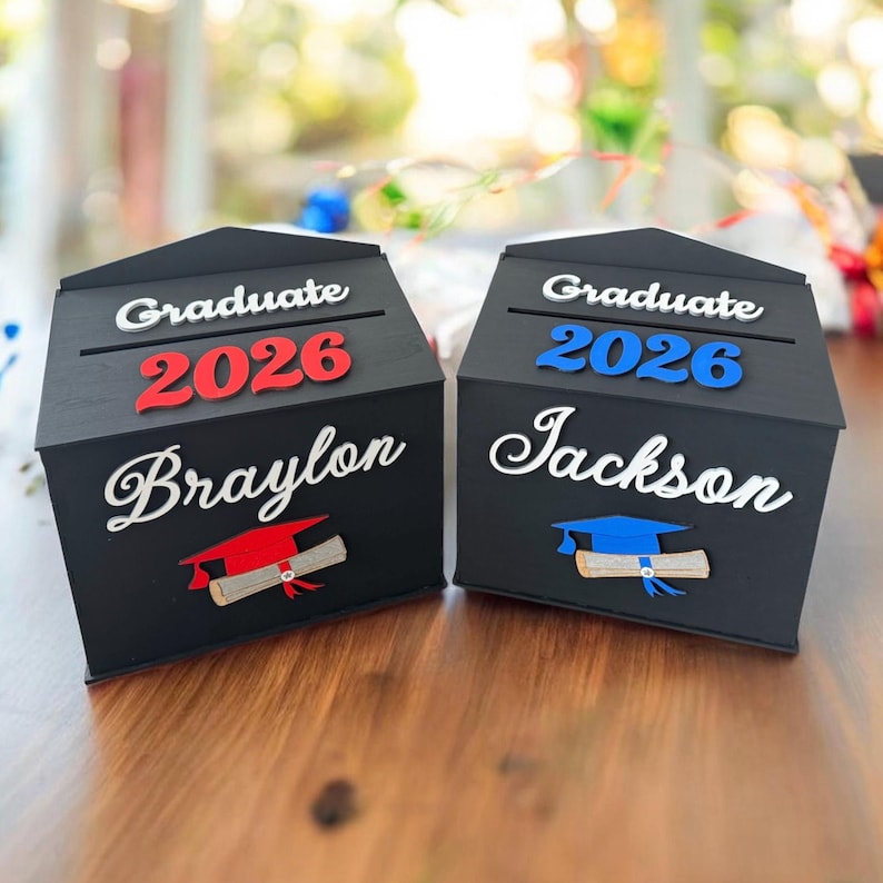 K&ouml;nnte beinhalten: Zwei schwarze Abschlussboxen aus Holz mit den W&ouml;rtern "Graduate 2026" in Wei&szlig; und Rot/Blau. Jede Box hat einen Namen in wei&szlig;er Schrift und eine Grafik mit Abschlusskappe und Diplom. Eine Box tr&auml;gt die Aufschrift "Braylon", die andere "Jackson".