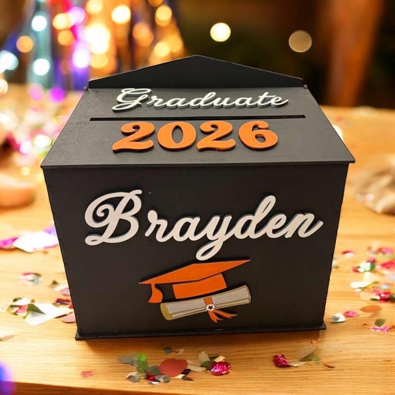 K&ouml;nnte beinhalten: Eine schwarze Abschlussbox mit dem Namen "Brayden" in wei&szlig;er Schrift. Die Box hat "Graduate 2026" in orangefarbenen Buchstaben. Eine Abschlusskappe und ein Diplom sind ebenfalls auf der Box abgebildet.