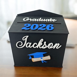 K&ouml;nnte beinhalten: Eine schwarze Holz-Abschlussbox mit dem Namen "Jackson" in wei&szlig;er Schrift. Das Jahr "2026" ist blau und das Wort "Graduate" ist silberfarben. Eine blaue Abschlusskappe und ein Diplom sind ebenfalls abgebildet.