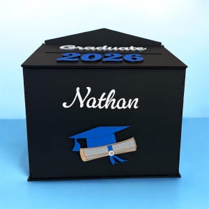 K&ouml;nnte beinhalten: Eine schwarze Abschlussbox mit dem Namen "Nathan" in wei&szlig;er Schrift. Die Box zeigt das Wort "Graduate" und das Jahr "2026" in Blau. Eine blaue Abschlusskappe und eine Urkunde sind ebenfalls auf der Box abgebildet.