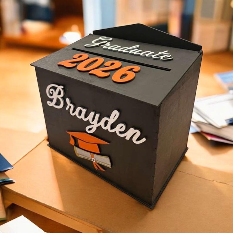 K&ouml;nnte beinhalten: Eine schwarze Holz-Abschlussbox mit dem Wort "Graduate" in wei&szlig;er Schrift &uuml;ber den orangefarbenen Zahlen "2026". Der Name "Brayden" steht in wei&szlig;er Schrift darunter, zusammen mit einer Grafik von Abschlusskappe und Diplom.