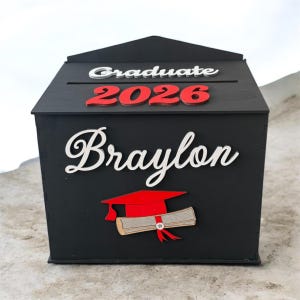 K&ouml;nnte beinhalten: Eine schwarze Holz-Abschlussbox mit den Worten "Graduate 2026" in Wei&szlig; und Rot. Der Name "Braylon" wird in wei&szlig;er Schrift angezeigt. Eine rote Abschlusskappe und ein Diplom sind ebenfalls abgebildet.