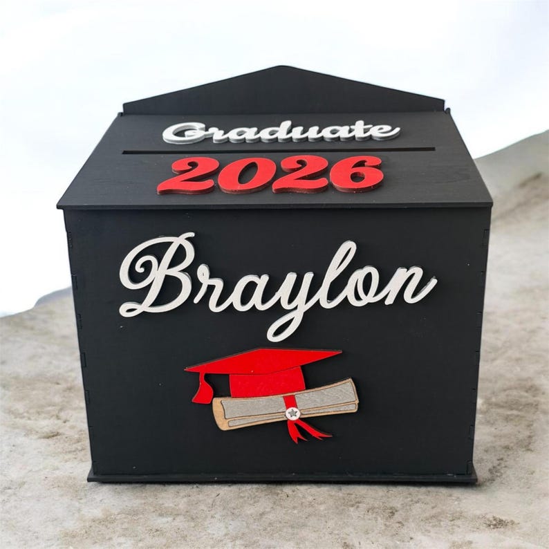 K&ouml;nnte beinhalten: Eine schwarze Holz-Abschlussbox mit den Worten "Graduate 2026" in Wei&szlig; und Rot. Der Name "Braylon" wird in wei&szlig;er Schrift angezeigt. Eine rote Abschlusskappe und ein Diplom sind ebenfalls abgebildet.