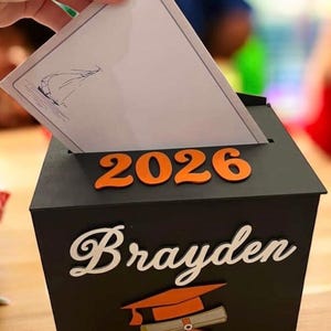 K&ouml;nnte beinhalten: Eine schwarze Abschlussbox mit dem Namen "Brayden" in wei&szlig;er Schrift und dem Jahr "2026" in Orange. Eine Abschlusskappe und ein Diplom sind ebenfalls abgebildet. Eine Hand steckt eine Karte in die Box.
