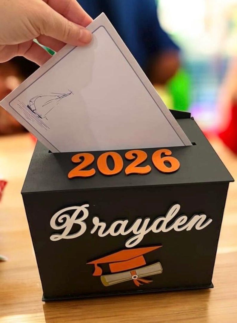 K&ouml;nnte beinhalten: Eine schwarze Abschlussbox mit dem Namen "Brayden" in wei&szlig;er Schrift und dem Jahr "2026" in Orange. Eine Abschlusskappe und ein Diplom sind ebenfalls abgebildet. Eine Hand steckt eine Karte in die Box.