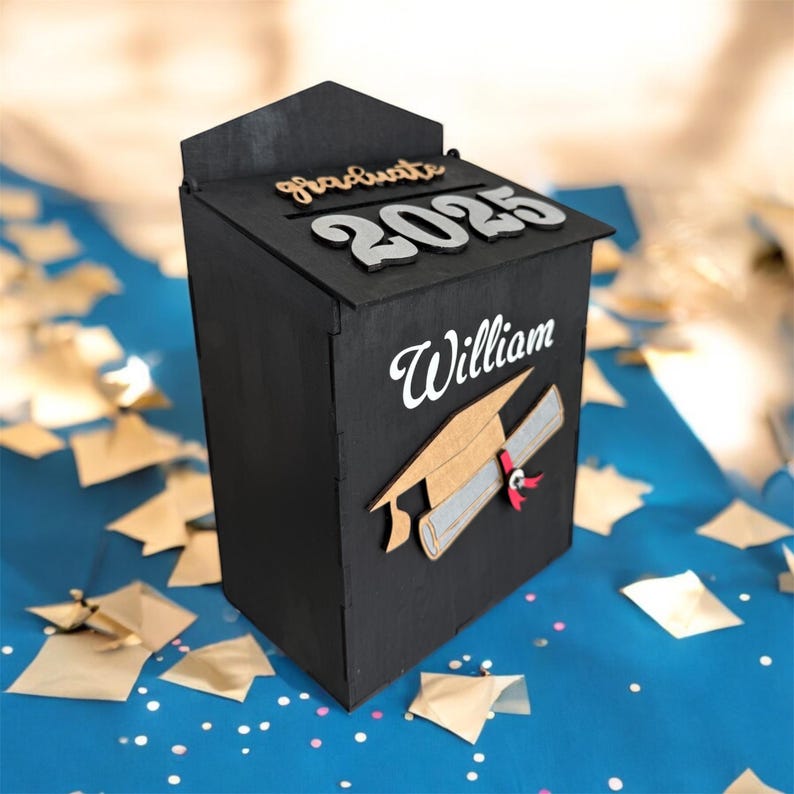 K&ouml;nnte beinhalten: Eine schwarze Holz-Abschlussbox mit dem Wort "Graduate" und dem Jahr "2025" oben. Der Name "William" ist auf der Vorderseite zusammen mit einer Abschlusskappe und einem Diplom abgebildet. Die Box ist mit goldenen und silbernen Akzenten verziert.