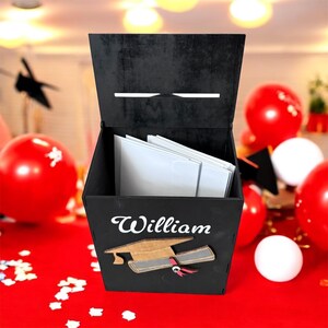 K&ouml;nnte beinhalten: Eine schwarze Holz-Abschlusskartenbox mit dem Namen "William" in wei&szlig;er Schrift. Eine Abschlusskappe und ein Diplom sind unter dem Namen abgebildet. Die Box ist ge&ouml;ffnet und zeigt wei&szlig;e Umschl&auml;ge im Inneren. Rote und wei&szlig;e Luftballons sind im Hintergrund.