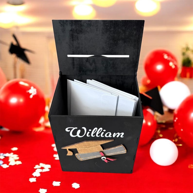 K&ouml;nnte beinhalten: Eine schwarze Holz-Abschlusskartenbox mit dem Namen "William" in wei&szlig;er Schrift. Eine Abschlusskappe und ein Diplom sind unter dem Namen abgebildet. Die Box ist ge&ouml;ffnet und zeigt wei&szlig;e Umschl&auml;ge im Inneren. Rote und wei&szlig;e Luftballons sind im Hintergrund.