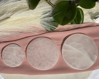 Könnte beinhalten: Drei weiße Selenit-Ladeplatten in verschiedenen Größen auf einer rosa Stoffoberfläche. Die Platten sind glatt und haben eine durchscheinende Optik.