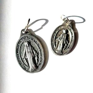 May include: Two silver-colored oval earrings. Each earring features an embossed image of a robed figure with outstretched arms, surrounded by text. The text includes the words "O Mary, Conceived Without Sin, Pray For Us Who Have Recourse To Thee."