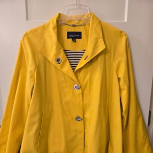 May include: A bright yellow jacket with a wide collar, long sleeves, and a front button closure featuring silver-toned hardware. The interior lining displays a black and white horizontal striped pattern. A Jones New York Signature label is visible.