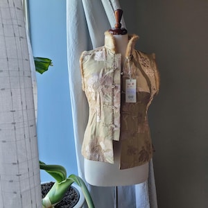 May include: A gold-colored vest with a floral pattern and fur trim. The vest has a mandarin collar and frog closures. A tag hangs from the vest.