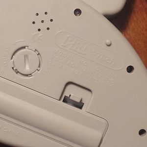 May include: Close-up of the back of a white electronic device. The device has a battery compartment with a screw-in cover and a compartment for two AA batteries. The text "PRO Tech" and "MODEL NO: GJ-705" are visible.