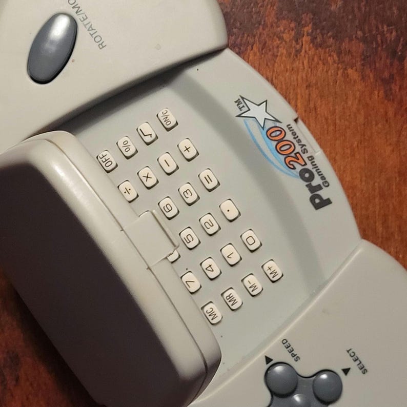 May include: A light gray Pro200 Gaming System handheld game console. It features a numeric keypad, function buttons, and directional controls. The console has the text "Pro200 Gaming System" and "ROTATE/MO" printed on it.
