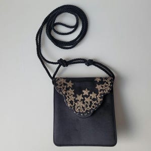 Pode incluir: Pequena bolsa transversal de cetim preto com aba decorativa. A aba é adornada com bordados florais dourados. A bolsa tem uma alça de corda preta e é exibida em um fundo branco.