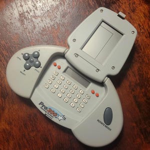 May include: A light gray Pro200 Gaming System, a handheld electronic device with a calculator keypad, a screen, and a directional pad. The device has buttons labeled "Level", "Select", and "Speed". The screen is open.