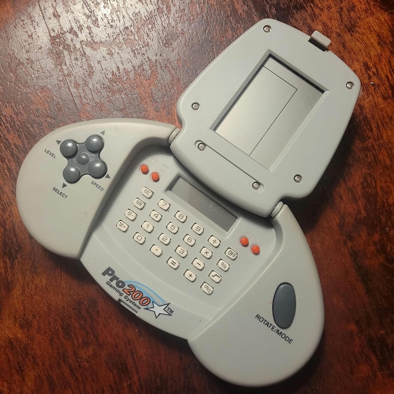 May include: A light gray Pro200 Gaming System, a handheld electronic device with a calculator keypad, a screen, and a directional pad. The device has buttons labeled "Level", "Select", and "Speed". The screen is open.
