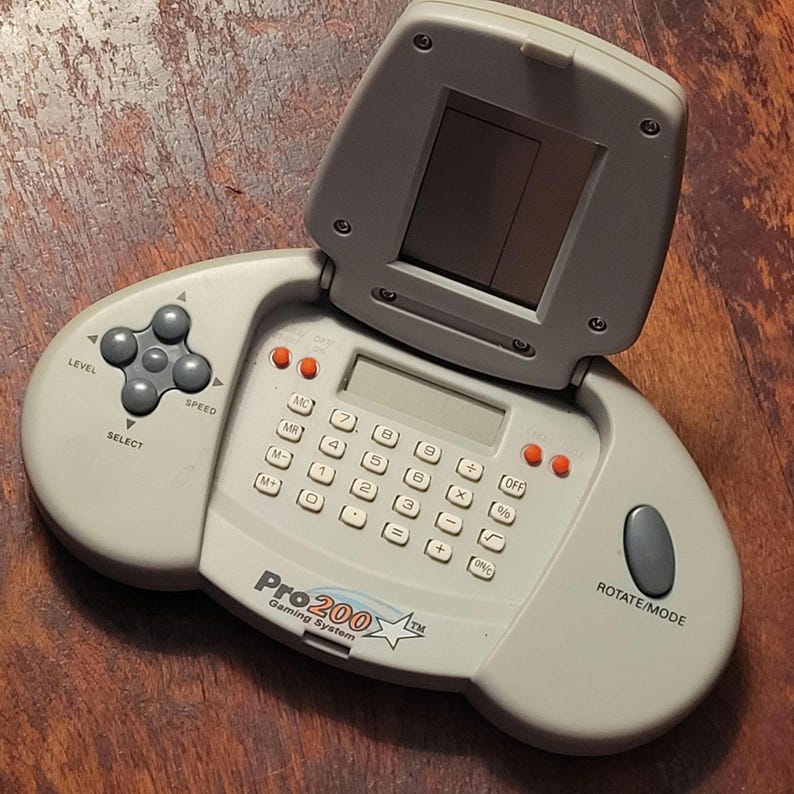 May include: A light gray Pro 200 Gaming System with a flip-up screen, calculator buttons, and a directional pad. The device has a gray and orange color scheme, with the text "Pro 200 Gaming System" visible.