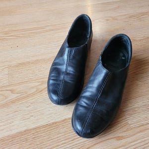 Puede incluir: Un par de zapatos sin cordones de cuero negro con punta redondeada, cuello bajo y una costura vertical prominente que recorre el centro del empeine. Los zapatos tienen un acabado liso y un dise&ntilde;o sencillo y funcional sin cordones ni hebillas.