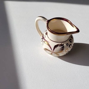 Puede incluir: Un pequeño creamer de cerámica blanca con un diseño floral dorado y burdeos. El creamer tiene un cuerpo redondeado y un pequeño pico.