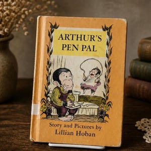 Puede incluir: Libro infantil vintage titulado "Arthur's Pen Pal" de Lillian Hoban. La portada es de color naranja descolorido con ilustraciones de un mono escribiendo y un niño saltando a la cuerda. El libro está ligeramente desgastado, lo que sugiere que es antiguo.
