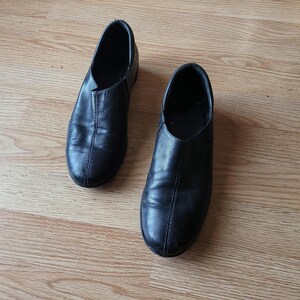 Puede incluir: Un par de zapatos negros sin cordones con un acabado liso, punta redondeada y una costura vertical prominente que recorre el centro del empeine. Estos zapatos de perfil bajo tienen un dise&ntilde;o sencillo y limpio sin cordones ni herrajes.