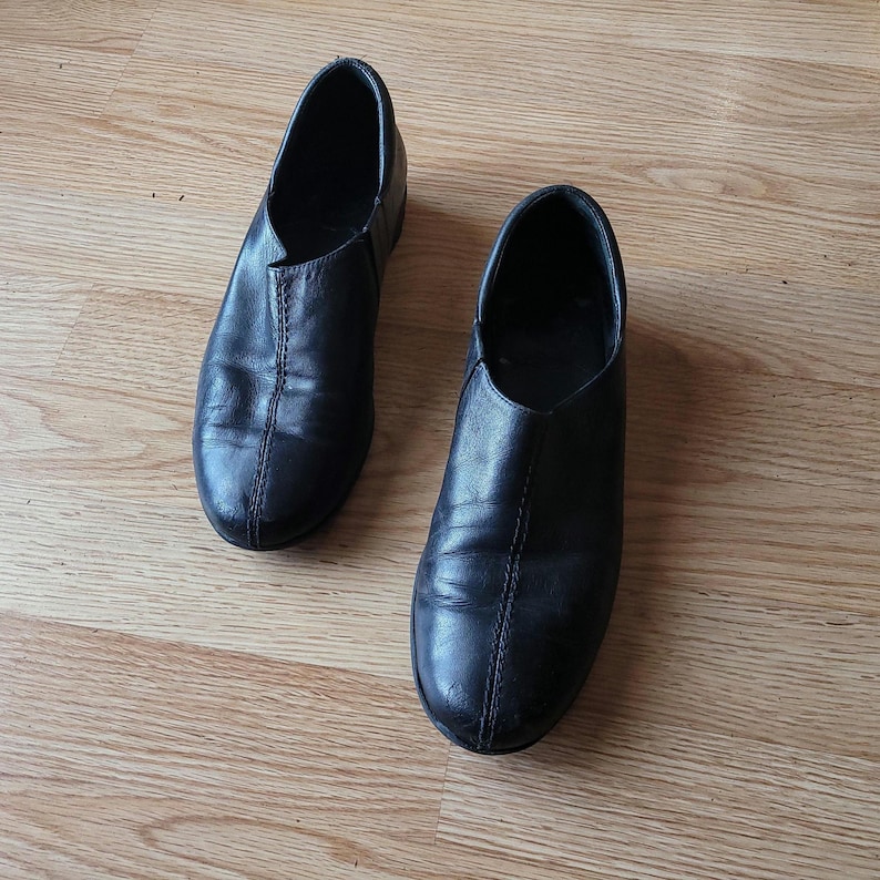 Puede incluir: Un par de zapatos negros sin cordones con un acabado liso, punta redondeada y una costura vertical prominente que recorre el centro del empeine. Estos zapatos de perfil bajo tienen un dise&ntilde;o sencillo y limpio sin cordones ni herrajes.
