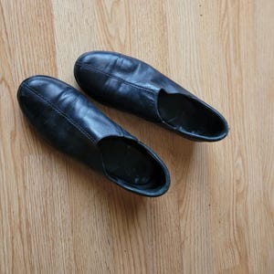 Puede incluir: Un par de zapatos sin cordones de cuero negro con punta redondeada, una costura central a lo largo del empeine y un cuello bajo. Los zapatos tienen un acabado liso, un dise&ntilde;o sencillo y sin adornos con suela plana y forro interior oscuro.