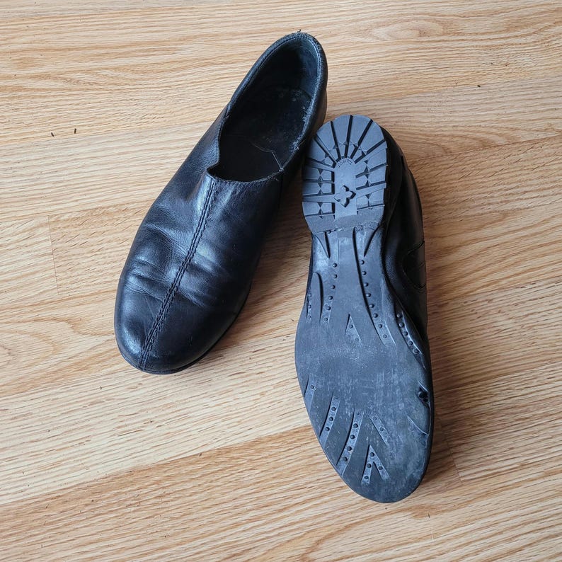 Puede incluir: Un par de zapatos negros sin cordones. Un zapato est&aacute; colocado para mostrar la parte superior de cuero liso con una costura central, mientras que el otro est&aacute; volteado para mostrar la suela de goma negra con un patr&oacute;n de relieve texturizado.