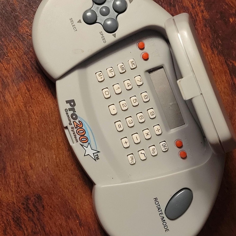 May include: A light gray Pro 200 Gaming System handheld game console. It features a calculator-style keypad with number and function buttons, a small screen, and a directional pad with select and speed buttons.