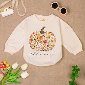 May include: Cream-colored baby bodysuit with a floral pumpkin design. The pumpkin is filled with colorful flowers and foliage. The name "Elliani" is written in script below the pumpkin. Sunglasses and a toy camera are in the background.