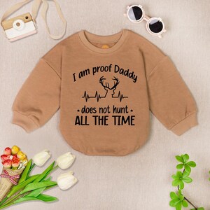 May include: Tan long-sleeve sweatshirt with a black graphic that reads "I am proof Daddy does not hunt all the time". The graphic also includes a deer silhouette and a heartbeat line.