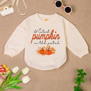 May include: A cream colored long-sleeved baby onesie with the text "Cutest pumpkin in the patch" in orange and black lettering. The onesie has a graphic of two orange pumpkins with green stems and leaves.