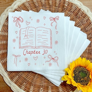 May include: White paper napkins with a red illustration of an open book, bows, hearts, and flowers. The text "Chapter 30" is below the book. The napkins are stacked on a woven placemat with a sunflower in the corner.