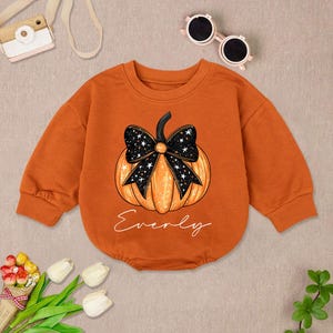 May include: Orange baby bodysuit with a pumpkin graphic and a black bow with white stars. The word "Everly" is written in cursive below the pumpkin. The long-sleeved bodysuit is made of a soft material.