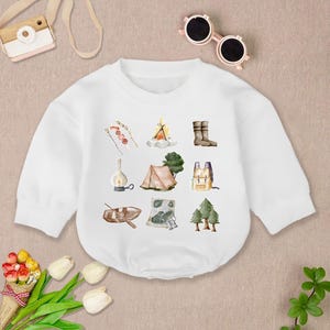 May include: White long-sleeved baby bodysuit with watercolor illustrations of camping gear. The design includes a campfire, tent, boots, backpack, and other outdoor elements. The bodysuit is made of soft fabric.