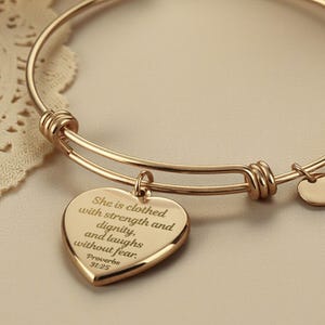 May include: A gold-colored bangle bracelet with a heart-shaped charm and two small round charms. The heart charm has the inscription "She is clothed with strength and dignity, and laughs without fear. Proverbs 31:25."