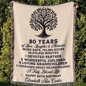 May include: A beige woven throw blanket with a black tree design and the text "80 Years of Love, Laughter & Memories 29,220 Days, 701,280 Hours 42,076,800 Minutes 1 Devoted Partner 5 Wonderful Children 6 Loving Grandchildren 3 Cherished Great-Grandchildren A Truly Blessed Life Happy 80th Birthday Elizabeth Ann Carter".