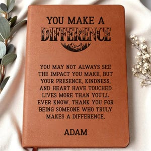 May include: A brown leather journal with the words "YOU MAKE A DIFFERENCE" in black. The journal has a mountain graphic and a heartfelt message. The name "ADAM" is printed at the bottom.
