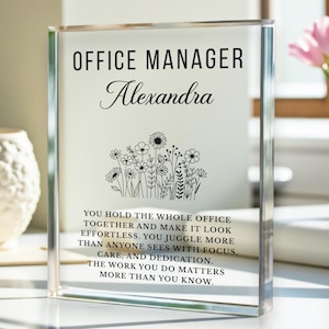 May include: A clear acrylic block with the words "OFFICE MANAGER Alexandra" in black. Below the text is a floral design. The block also includes an inspirational message about the office manager's work.