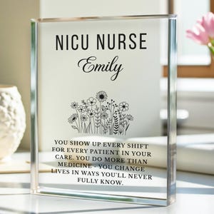 May include: A clear acrylic block with the words "NICU NURSE Emily" in black. Below the name is a floral design. The text reads: "YOU SHOW UP EVERY SHIFT FOR EVERY PATIENT IN YOUR CARE. YOU DO MORE THAN MEDICINE - YOU CHANGE LIVES IN WAYS YOU'LL NEVER FULLY KNOW."