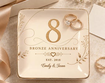 May include: A square cream-colored ceramic trinket dish with gold trim featuring the number 8, the text BRONZE ANNIVERSARY, EST. 2018, and the names Emily & Jason in gold lettering. Two gold rings are resting on the dish, and one ring is nearby.