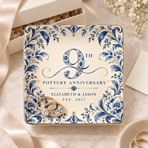 May include: A square ceramic trinket dish with a cream background and blue floral border. The center features the text 9TH POTTERY ANNIVERSARY, ELIZABETH & JASON, and EST. 2017. Two gold-toned rings with clear stones rest on the dish surface.