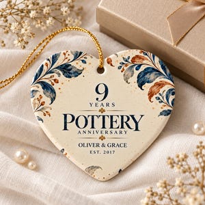 May include: A heart-shaped ceramic ornament with a cream background, featuring blue and copper-toned floral illustrations. The text reads 9 YEARS POTTERY ANNIVERSARY, OLIVER & GRACE, EST. 2017. It includes a gold hanging cord.
