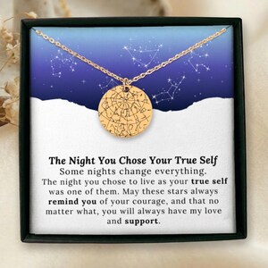 May include: A gold-colored necklace with a round pendant featuring a star map design. The necklace is displayed in a black box with a blue and white background. Text on the box reads: "The Night You Chose Your True Self."