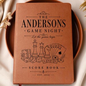 May include: A tan leather-bound score book featuring black engraved text and illustrations. The cover displays the text THE ANDERSONS GAME NIGHT Let the games begin SCORE BOOK EST. 2026, alongside graphics of dominoes, chess pieces, playing cards, and dice.